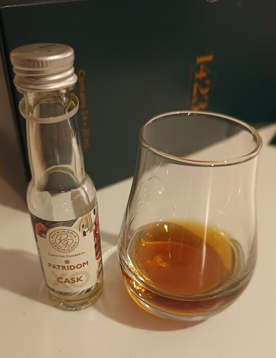 Miniflaska med Patridom Cask-rom bredvid ett glas med gyllene rom på ett vitt bord.