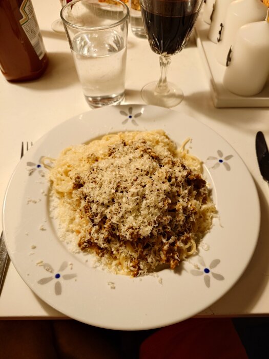 Spaghetti med köttfärssås toppad med riven parmesan på en vit tallrik, omgiven av ett glas vin och vatten.