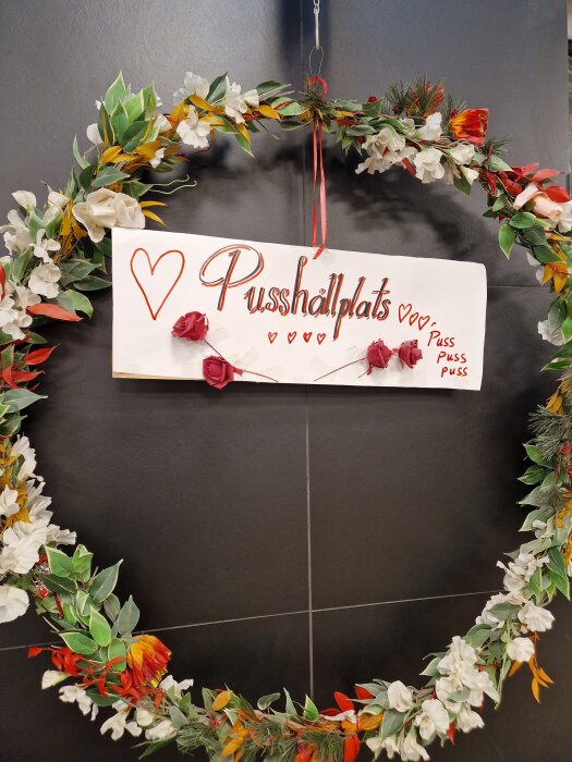 Krans av blommor och blad med en skylt som säger "Pusshållplats" dekorerad med hjärtan och texten "Puss puss puss" på en mörk vägg.