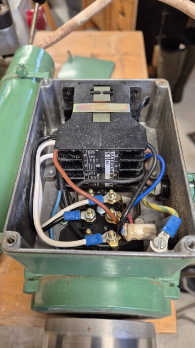 Närbild av en elektrisk kontaktor med synliga ledningar och anslutningar, monterad i en grön metallhölje, med bakgrund av en verkstadsmiljö.