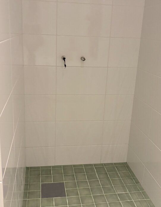 Duschutrymme med vita väggar, stora kakelplattor och en golvyta med gröna klinkerplattor. Notera avloppsbrunn och kakelremsor i duschen.