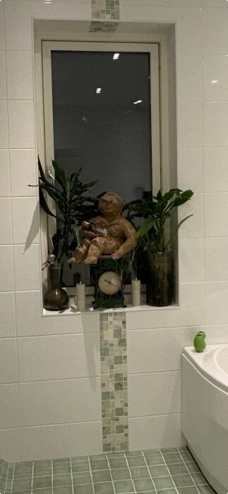 Fönsternisch i badrum med vita kakelplattor, gröna mosaikdetaljer och prydnader som växter och en staty. Gröna klinkerplattor på golvet.