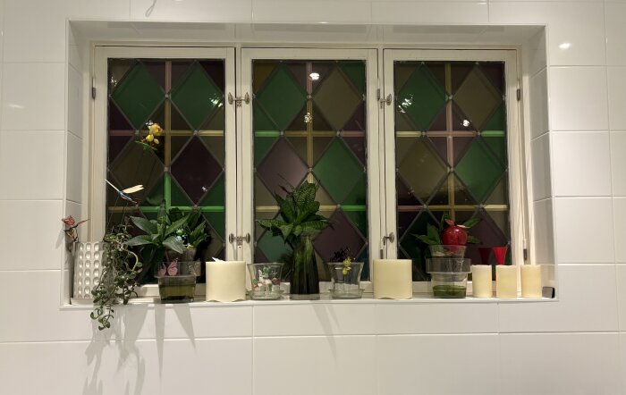 Fönster med färgat glas i rutmönster och vita kakelväggar. Balkong med växter och ljusdekorationer i badrumsmiljö.