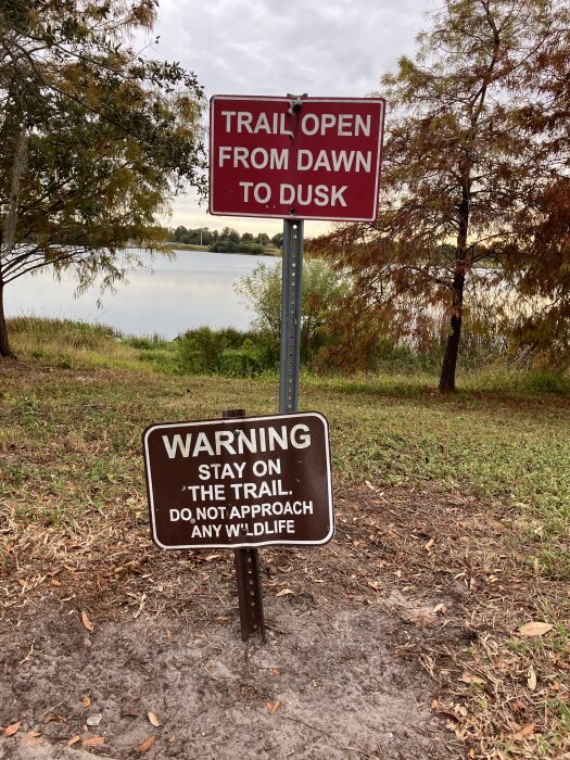 Två skyltar vid en stig nära en sjö, en visar "Trail open from dawn to dusk" och den andra varnar för att hålla sig på stigen och undvika vilda djur.
