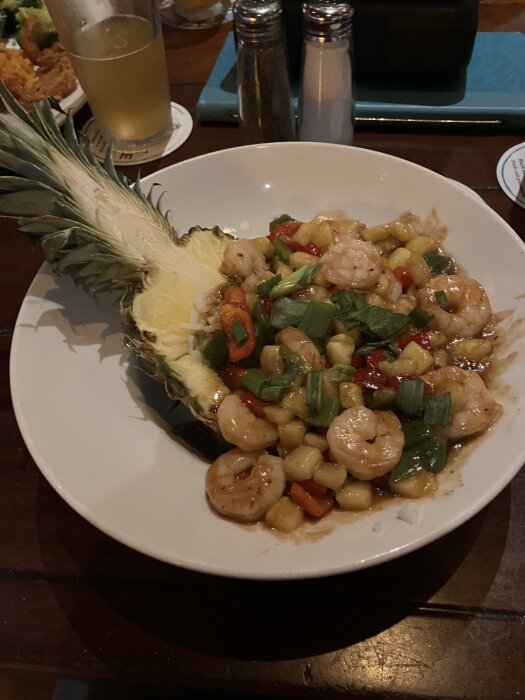 Ananas fylld med räkor, ananasbitar, paprika och grönsaker, serverad på en tallrik med en öl och kryddkvarnar i bakgrunden.