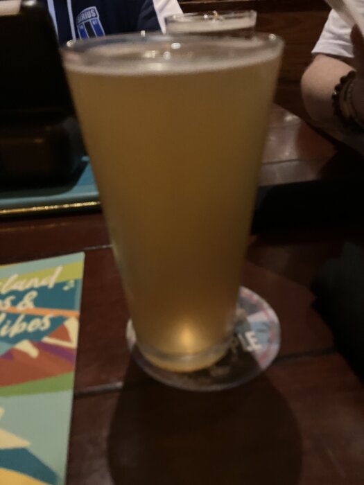 Ett glas med ljust öl på ett bord i en bar med en person i bakgrunden, samt en färgglad meny bredvid.