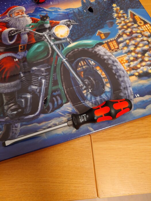 Julkalender med tomte på motorcykel och svart katt i snöig by. Skruvmejsel ovanpå kalendern på ett träbord.