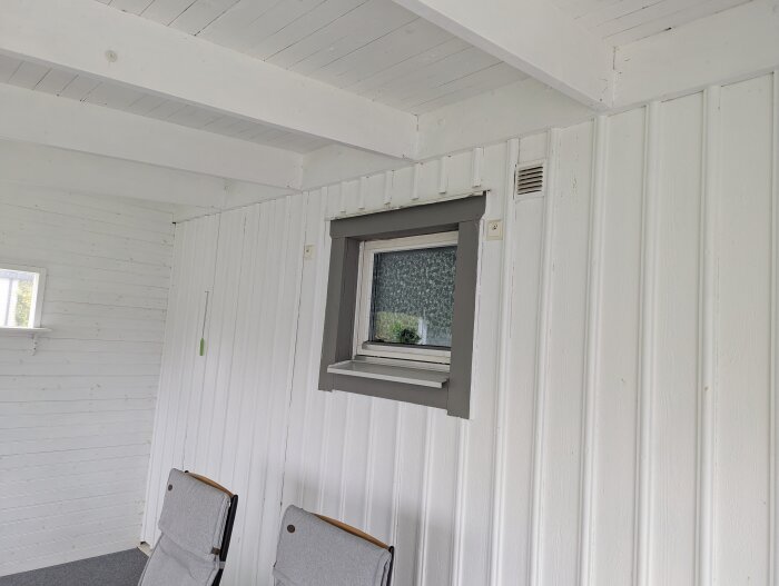 Vitmålad altan med ett fönster och en ventilationsöppning bredvid. Två stolar ses nedanför fönstret.