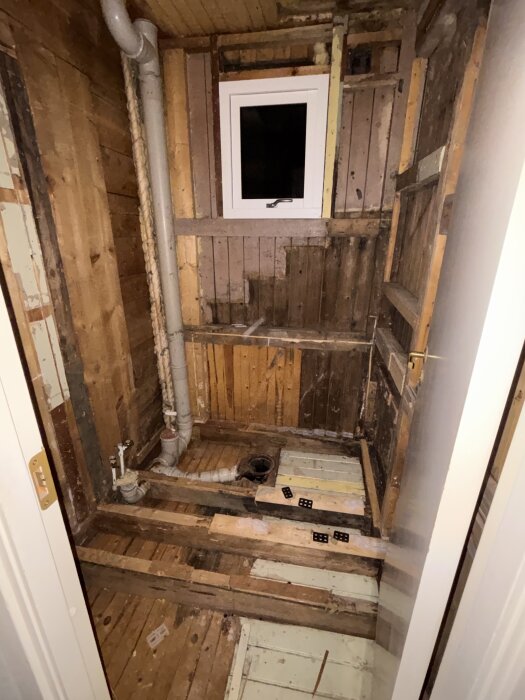 Renovering pågår i en liten tvättstuga med blottlagda träväggar och rör. Fönster ovanför och synlig golvstruktur.