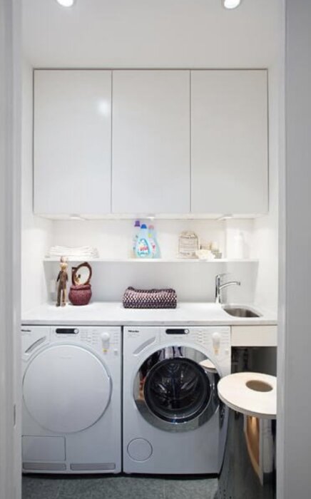 Liten tvättstuga med vit inredning, tvättmaskin och torktumlare sida vid sida, infälld diskho, överskåp och hyllor med tvättillbehör.