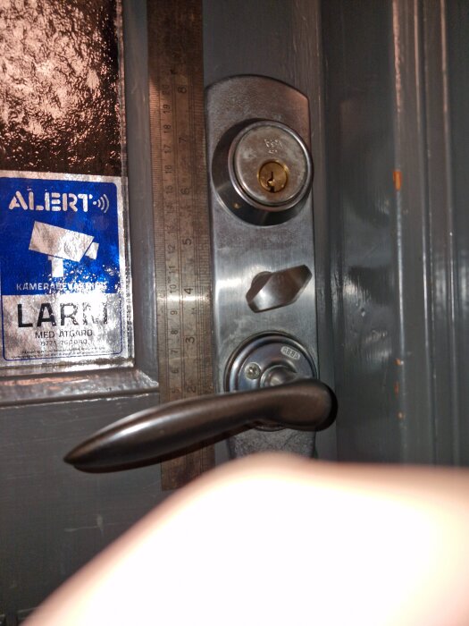 Dörrhandtag och låscylinder på grå ytterdörr, med linjal för att mäta avstånd.