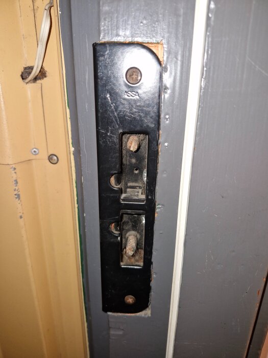 Närbild på en dörrkarm med ett svart ASSA-lås monterat, synliga träkanter vid låskistan och ett kilformat fall.