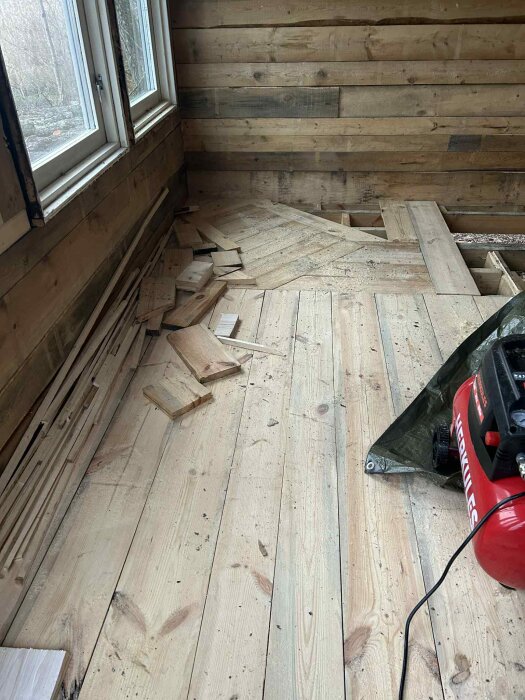 Träpaneler och bräder i en bastu under renovering, furuplankor på golvet, flera brädbitar staplade, och en kompressor i hörnet.