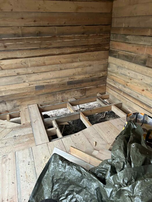 Halvbyggd bastu med grånade träväggar och delvis lagt furugolv. Verktyg och presenning syns på golvet.