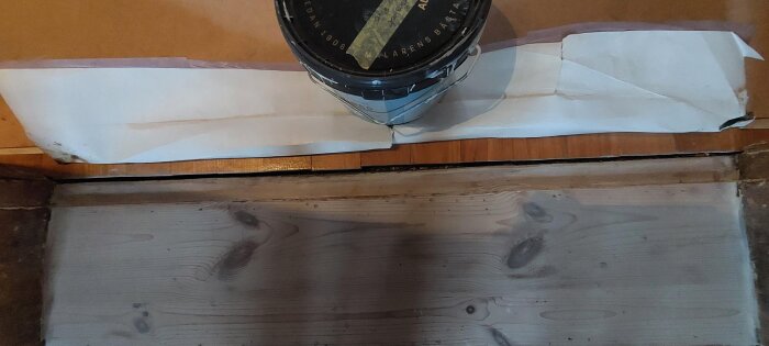 Sliten tröskel i sekelskiftesstil med ojämn yta bredvid nyare parkettgolv och en målarburk på papper ovanför.