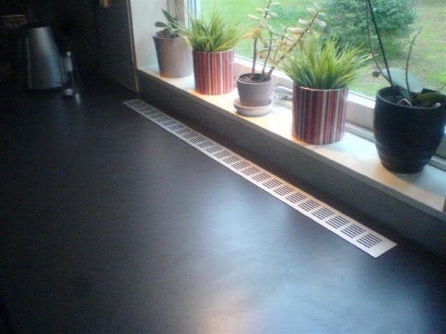 Svart bänkskiva med ventilationsgaller nära fönster, flera krukväxter på fönsterbrädan.