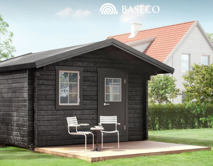 Svart friggebod med taköverhäng, två fönster och en dörr, omgiven av träd och en gräsmatta, med uteplats och stolar framför.