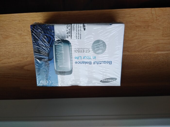 Förpackning av en Samsung-mobiltelefon, inplastad och placerad upp och ned på ett träbord.
