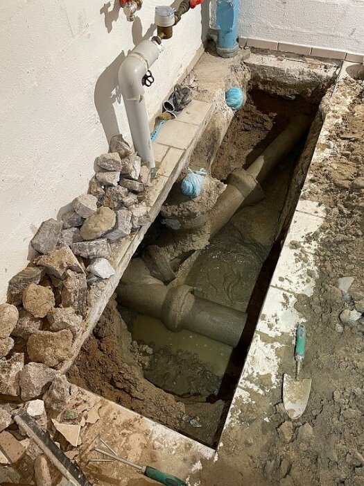 Källarrenovering med öppnad golvsektion visar avloppsrör och grävt hål, omgiven av uppgrävd jord och verktyg.