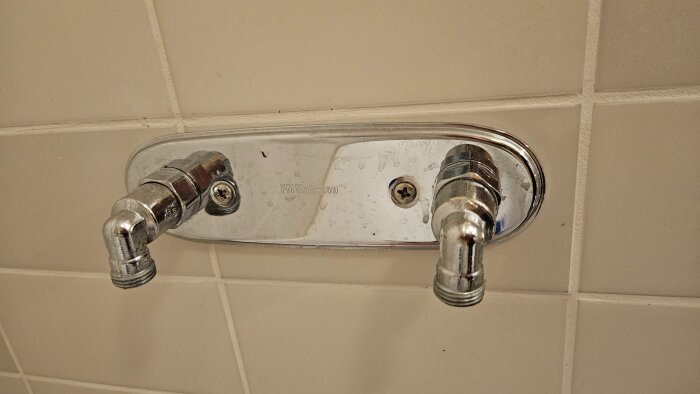 Kromad vattenrörsanslutning med två utgångar på en vit kakelvägg, märkt "FM Mattsson".