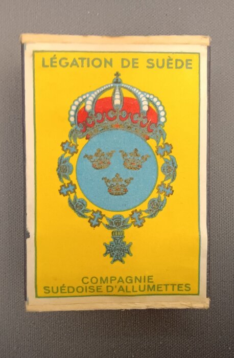 Svensk tändsticksask med motiv från svenska legationen, gul bakgrund, tre kronor, och text "Compagnie Suédoise d'Allumettes".