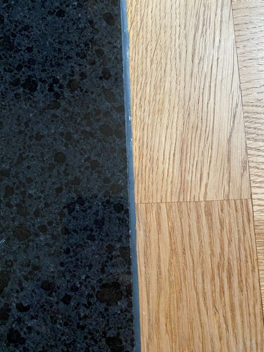 Svart granit möter ljus stavparkett med en svart mjukfog i övergången vid entrén, visar en rektangulär installation.