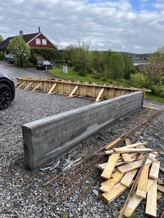 Betongmur under konstruktion med träformar och armeringsjärn på en uppfart, omgiven av gröna buskar och ett rött hus i bakgrunden.