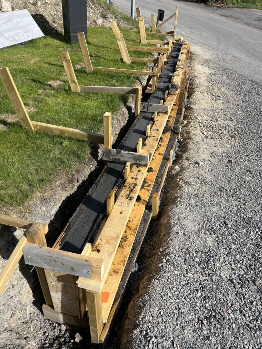 Betonggjutform vid en vägkant, med nygjuten betong i form av en mur, stöttad av träbjälkar och omgiven av grus.