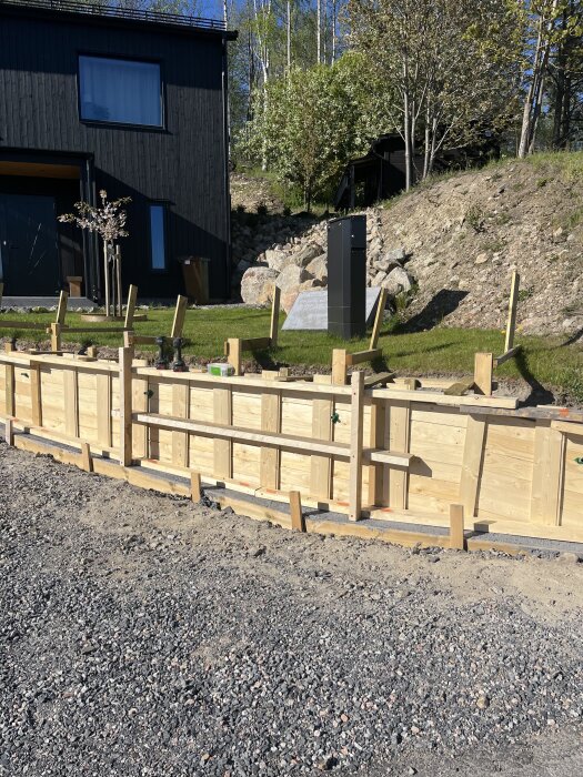 Form för gjutning av mur på en sluttande tomt framför ett svart hus, med träd och stenmur i bakgrunden.