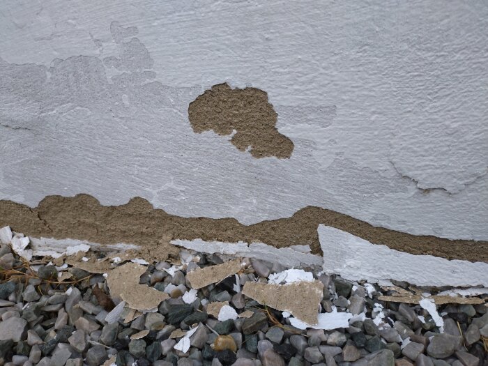 Putslagret och färgen har flagnat från nederdelen av en mur, med stenar liggande på marken nedanför.