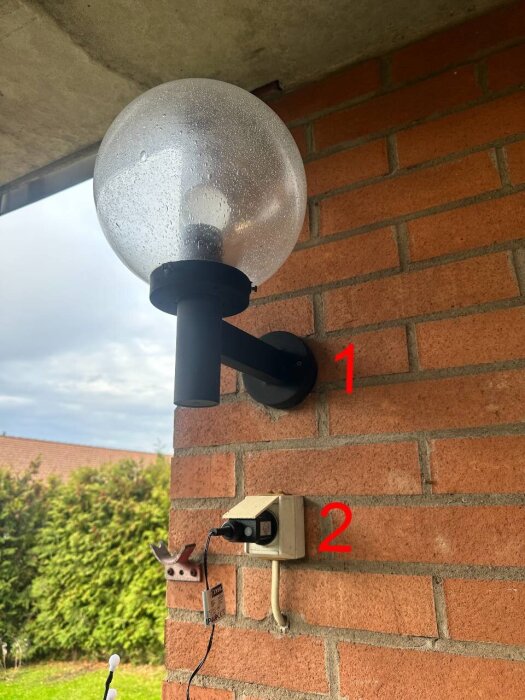 Väggmonterad utomhuslampa ovanför eluttag på tegelvägg, markerade med siffrorna 1 och 2.
