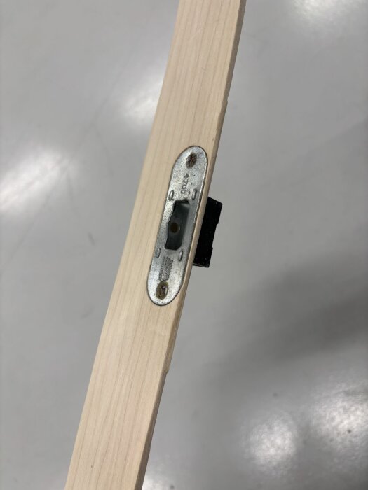 Dörrkarm av trä med metallplatta och låsdel monterad, fotograferad mot en ljus bakgrund.