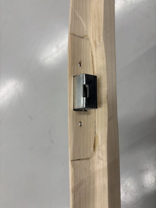 Trädörramas kant med ett metalliskt låsbleck infällt, omgiven av bearbetat trä med borrhål synliga.