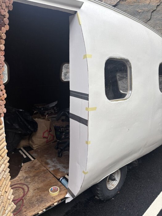 Mall i wellpapp för dörrkarmens kurvatur fasttejpad vid sidan av en vit husvagn med synliga hjul och kringliggande byggmaterial.