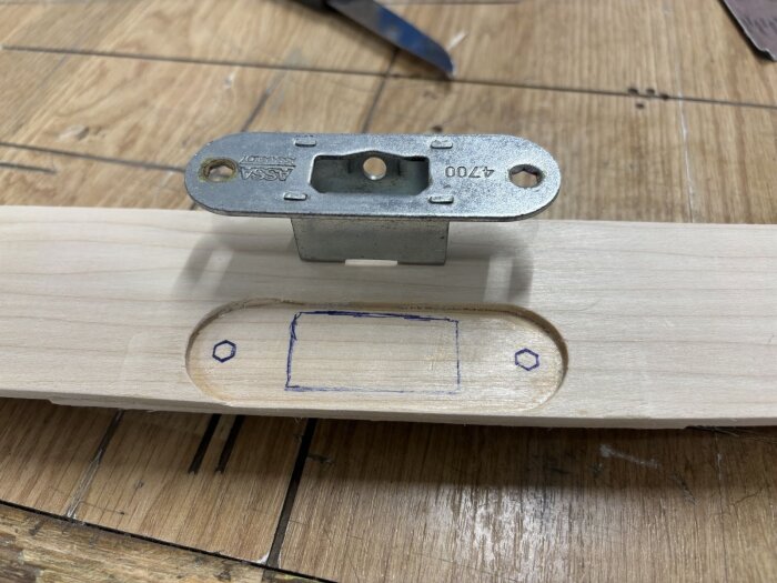 Infräst dörrlås i träbräda, med utmarkerade hål för skruvar och låskolv. Låset är från Assa och placeras i en urfräst del av träet.