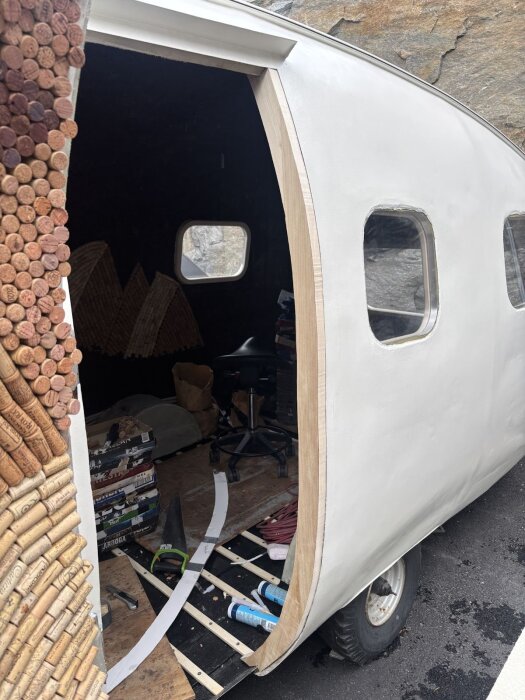 Hemmagjord dörrkarm under konstruktion på vit vagn, omgivna av verktyg som såg och limtuber; insidan har oordnade föremål och korkdetaljer runt dörröppningen.