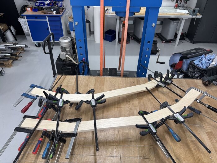 Träbitar som formas och limmas ihop med hjälp av klämmor, placerade på ett arbetsbord i en verkstad med blå och svarta verktygsskåp i bakgrunden.