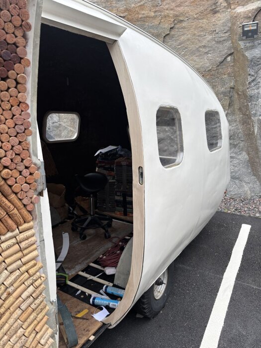 Vit husvagnskaross under renovering med öppen dörr och synliga träkarmar. Inuti syns en svart stol och diverse verktyg.