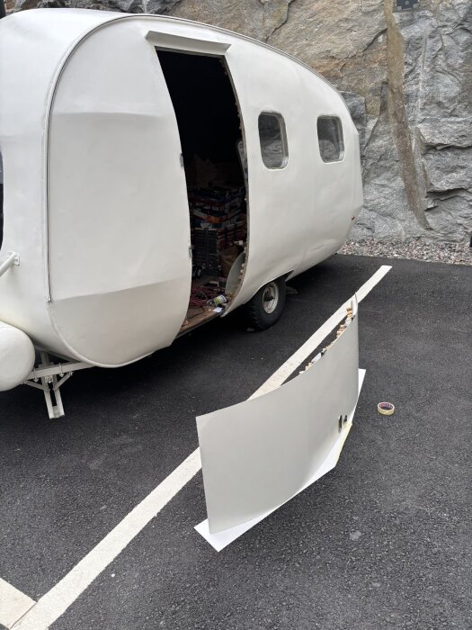 Vit husvagn med borttagen dörr placerad på marken framför, på en asfalterad yta vid en klippvägg.