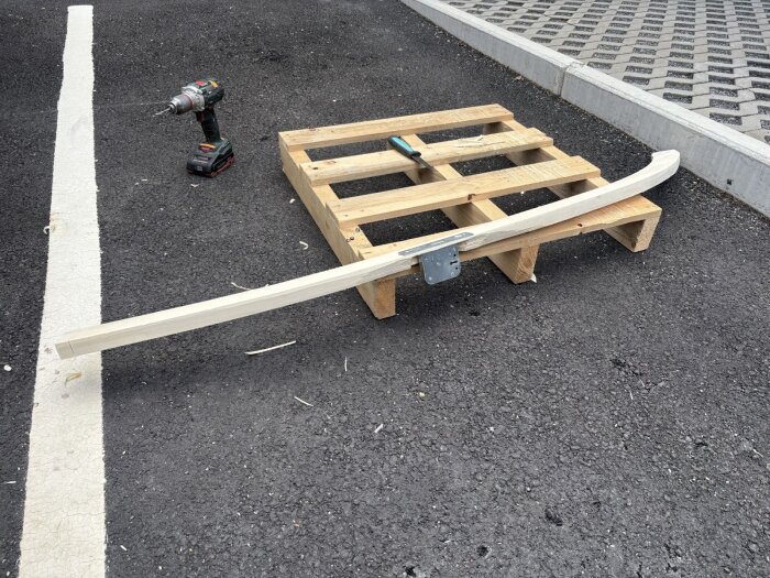 Träbit monterad på pall med metallbeslag, borrmaskin i bakgrunden på asfalterad yta.