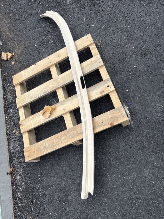 Trästycken formade i bågar för dörrkarm, ligger på en lastpall, med ett urtag för låsbleck, på en asfaltbelagd yta.