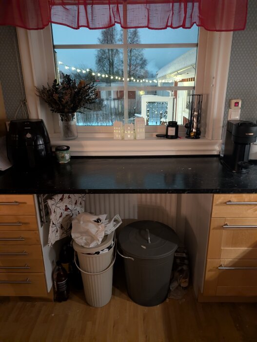 Fönster med röd gardin, utsikt över snötäckt trädgård och ljusslingor. Svart bänkskiva med hushållsartiklar, två soptunnor undertill.