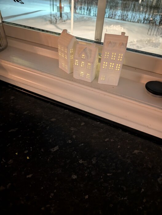 Miniatyrhus i vitt på en fönsterbräda med vinterlandskap utanför fönstret.