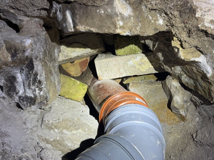 Rörinstallation i ett hål i en grundmur, omgiven av stenar och tegel som trycker mot röret från sidan.