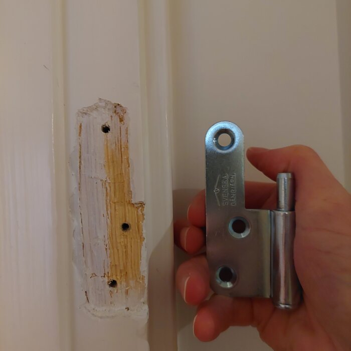 Hand håller gångjärn vid urtag i trädörrkarm, flera hål synliga.
