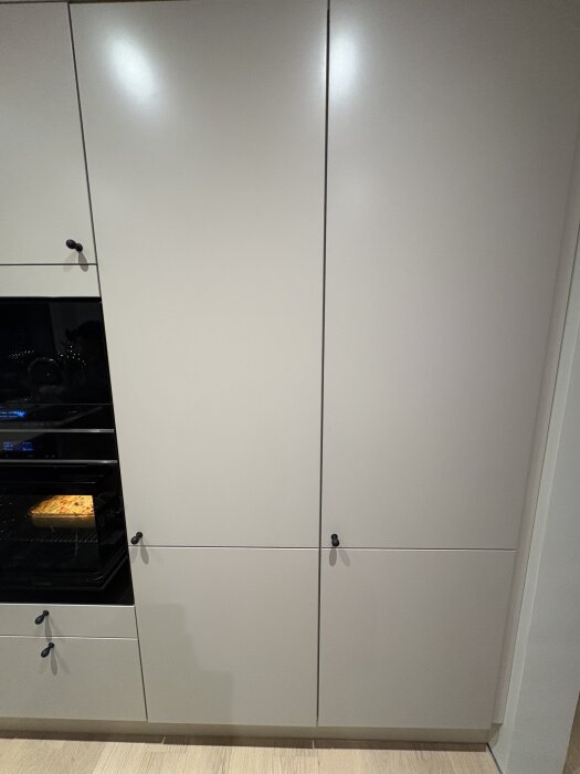 Grått köksskåp med Siemens kylskåp inbyggt bakom dörrpanel, bredvid ugn med mat som bakas inuti.