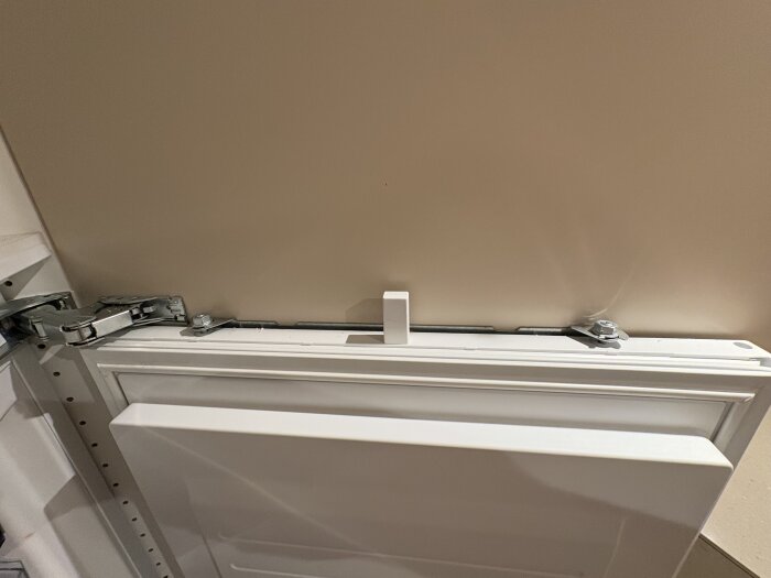 Justerbar fästanordning på Siemens kylskåp, fäst centrerat i dörrbladet, med gångjärn och monteringsdetaljer synliga mot en ljus vägg.