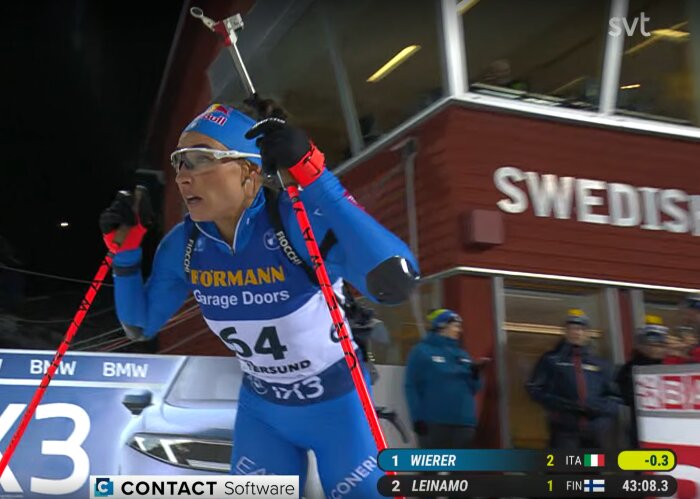 En tävlande skidskytt i blå dräkt med stavar passerar förbi ett rött hus med texten "SWEDISH", omgiven av åskådare och reklamskyltar.