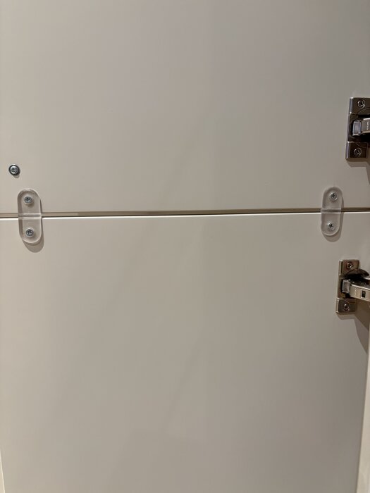 Transparent plastanordning med distans för att sätta rätt avstånd mellan dörrblad och kylskåpsdörr, synligt på vit yta med skruv och gångjärn.