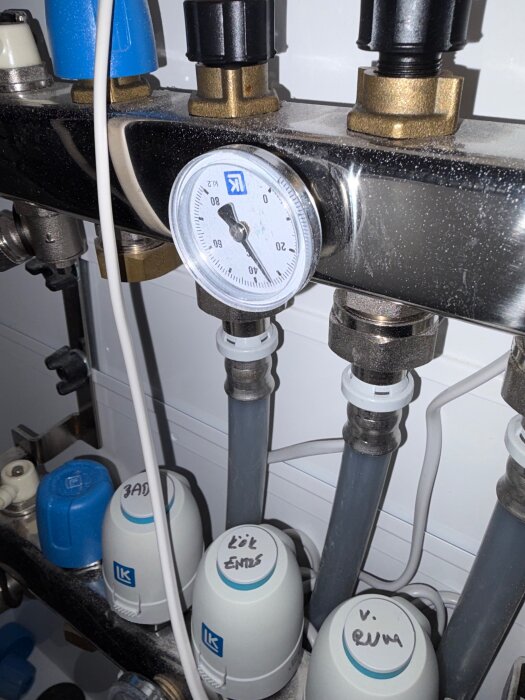 Manometer och rörkopplingar i golvvärmesystem, märkta termostater för badrum, kök och vardagsrum, fokus på temperaturfördelning.